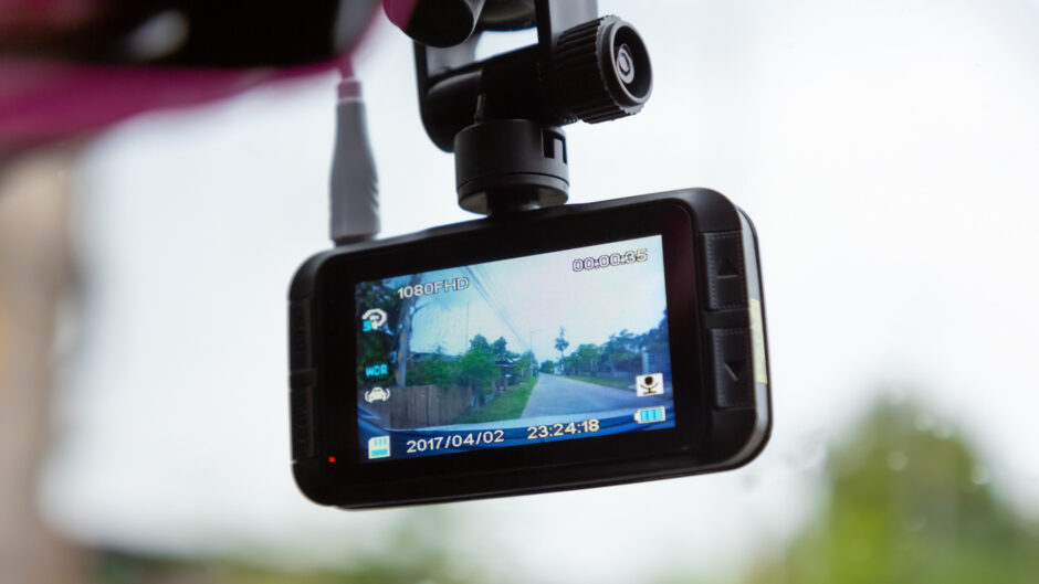 この画像は、車のフロントガラスに取り付けられたドライブレコーダーのカメラの写真になります。ドライブレコーダーのカメラは黒色で、左上隅に小さなカメラレンズがあります。ダッシュカメラには小さなLCDスクリーンがあり、道路と木々が並ぶ映像が表示されています。